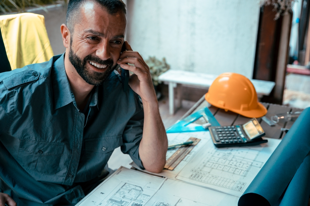 Bearded-Male-architect-portrait-using-phone-while-working-on-his-project-at-workplace